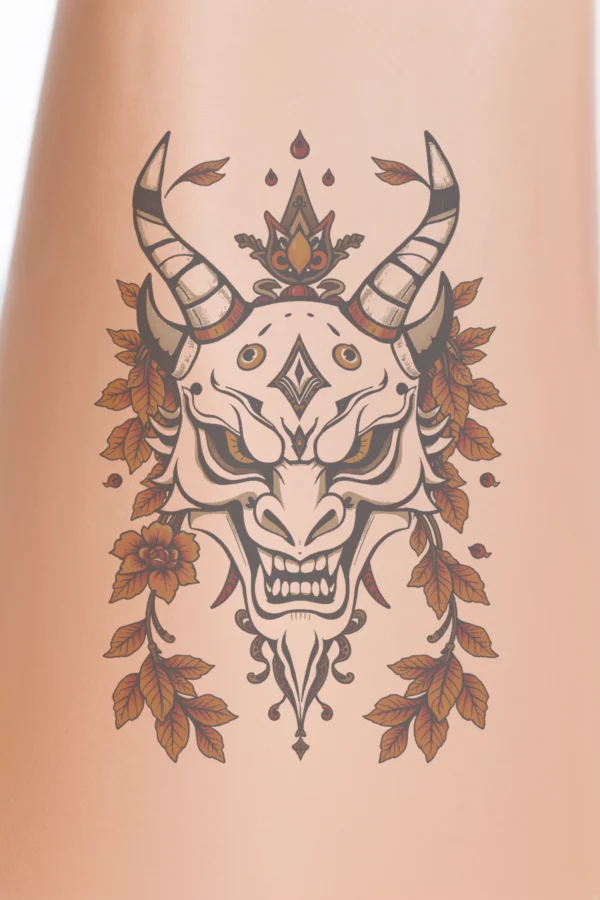 Close-up of a person's calf showcasing a temporary temporary tattoo of a stylized mask inspired by East Asian art. The predominantly black and white mask features a pointed nose, wide menacing grin, striking orange eyes, and a deep red mouth. Adorned