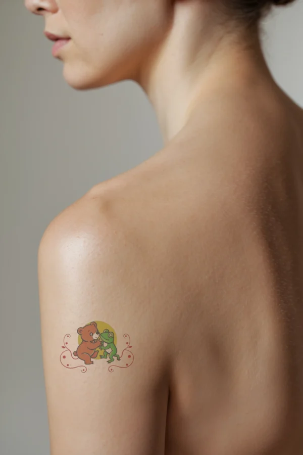 A temporary temporary tattoo on a person's shoulder features a colorful illustration of a friendly bear and frog. The bear, with reddish-brown fur, sits on its hind legs facing the green frog, which stands upright with yellow-green legs. Both animals are centered against