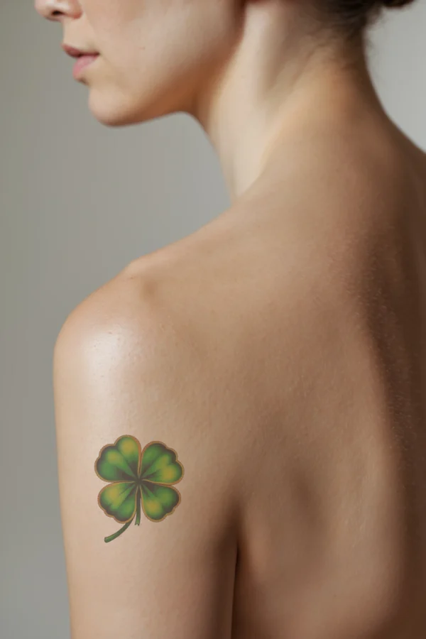 A vibrant temporary temporary tattoo of a four-leaf clover on a person's shoulder, depicted in green and orange hues. The clover's four symmetrical leaves are prominently displayed against a stark white background, highlighting its detail and contrasting colors. No other elements are